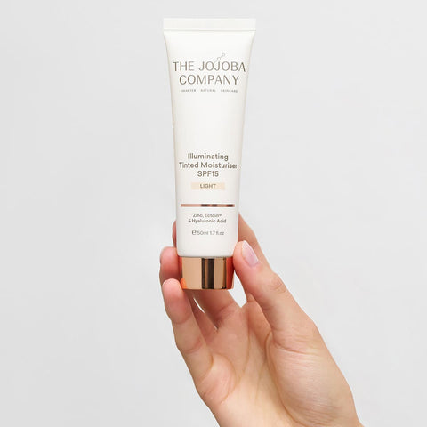 The Jojoba Company Illuminating Tinted Moisturiser SPF15 Light 50ml lifestyle shot of a hand holding the cream tube against a grey backdrop