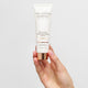 The Jojoba Company Illuminating Tinted Moisturiser SPF15 Light 50ml lifestyle shot of a hand holding the cream tube against a grey backdrop