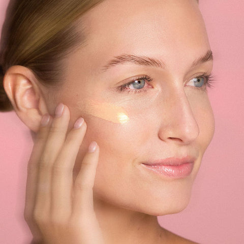 Woman applying a product to her face with a pink background.