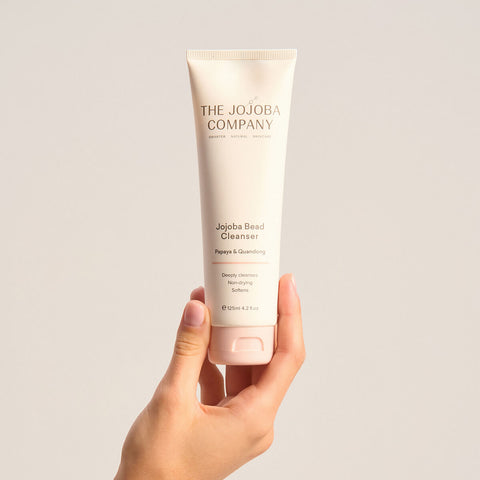The Jojoba Company Jojoba Bead Cleanser 125ml lifestyle shot of a hand holding the cream tube against a soft beige backdrop, front view of the tube