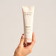 The Jojoba Company Jojoba Bead Cleanser 125ml lifestyle shot of a hand holding the cream tube against a soft beige backdrop, front view of the tube