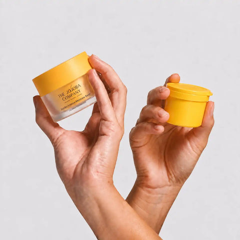 Two hands holding cosmetic jars against a light gray background. One hand holds a larger frosted jar with a yellow lid, and the other hand holds a smaller matching yellow jar.