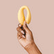 Hand holding The Natural Love Company Cistus, a remote controlled (remote included) U-shaped vibrator with dual vibrating motors (one at each end), it's crafted with a recycled ocean plastic internal body dressed in a buttery smooth silicone casing, which comes in a creamy yellow hue.