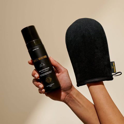 Hand holding a black self-tan spray can and black mitt against a beige background.