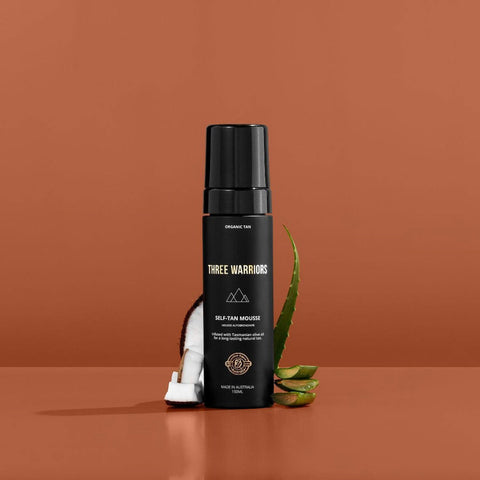 Three Warriors Self-Tan Mousse styled with coconut pieces and aloe vera leaves against an orange background.