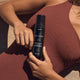 Close up of model laying on the beach in a red-brown swimsuit holding three warriors self tan mousse.