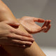 Hand applying a pinkish cream to leg against a neutral background.