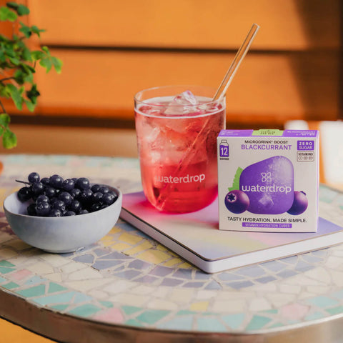 waterdrop Microdrink Boost Blackcurrant 12 servings lifestyle shot of the box, a glass of pink drink and a bowl of blueberries on an outdoor table