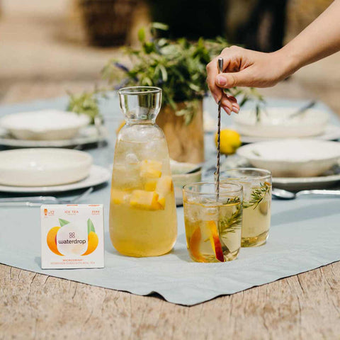 Person stirring a glass of iced tea with peach slices on a table with 'waterdrop' tea packaging.