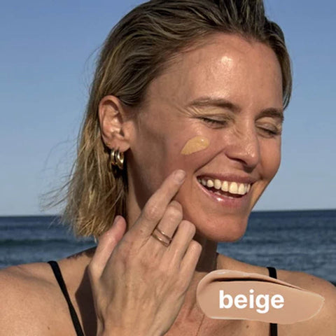 Woman applying beige sunscreen on her face with ocean in the background